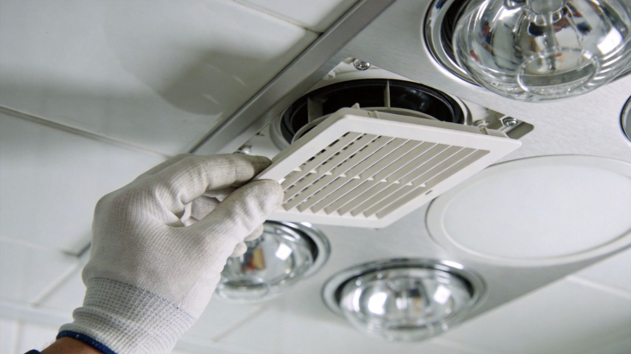 En person iført arbejdshandsker installerer eller vedligeholder et ventilationsgitter i loftet på et badeværelse.