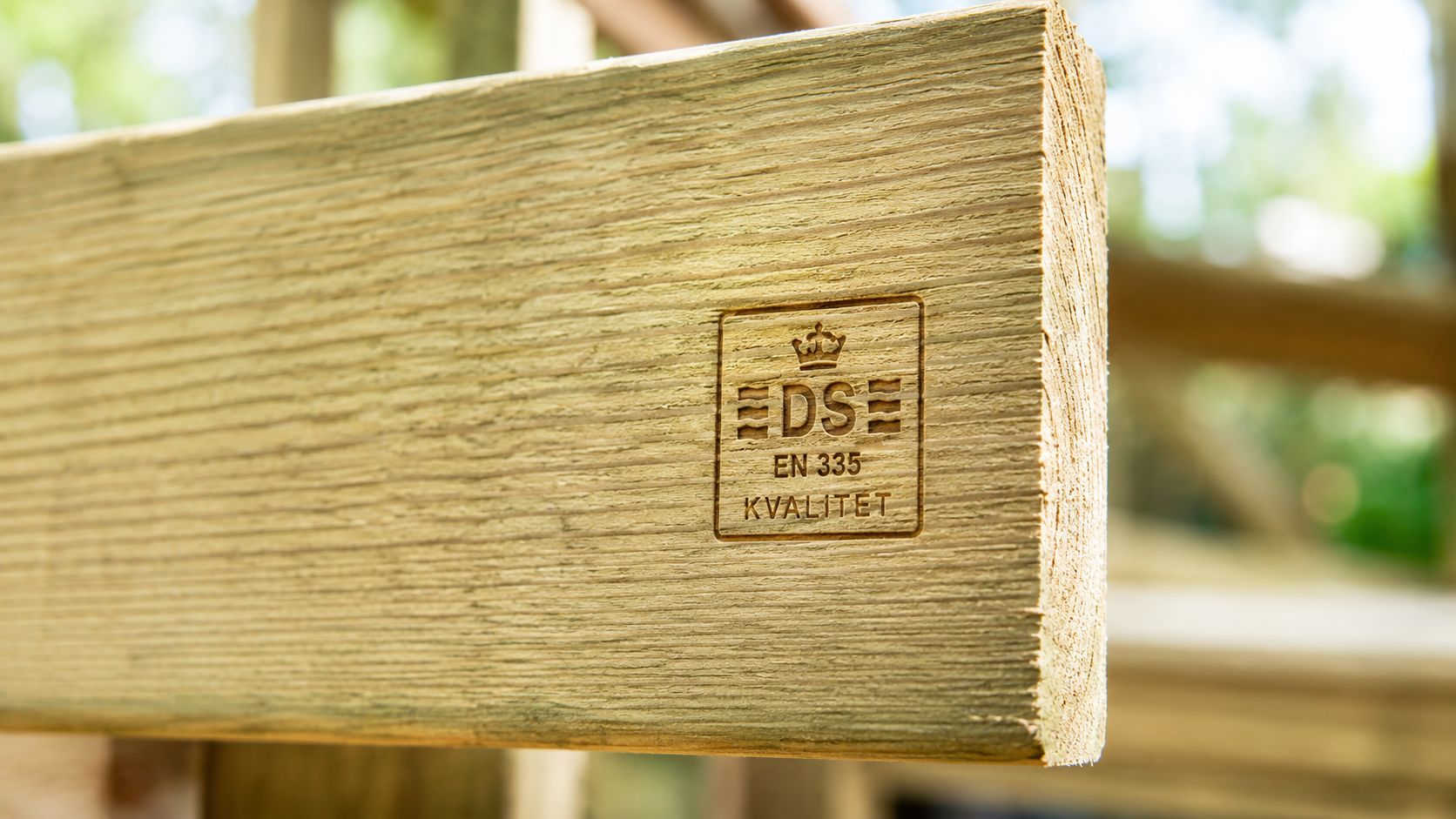 Nærbillede af en imprægneret træbjælke med et DS-kvalitetsstempel og EN 335 godkendelse for holdbarhed udendørs.